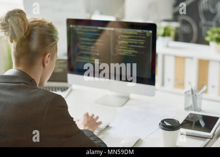 Back view portrait of contemporary young man coding at modern computer sitting at desk and working in startup project, shot from behind glass wall, co Stock Photo