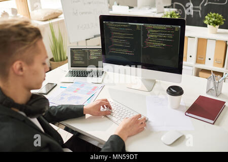 Portrait of contemporary young man coding at modern computer sitting at desk and working in startup project, focus on computer screen, copy space Stock Photo