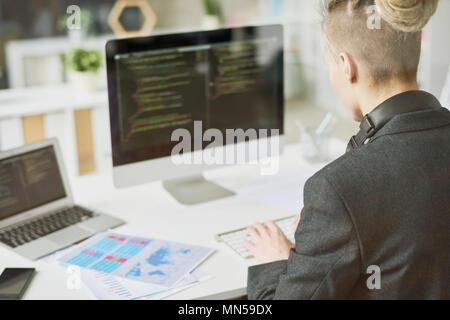 Over the shoulder view of contemporary young man coding at modern computer sitting at desk and working in startup project, shot from behind glass wall Stock Photo