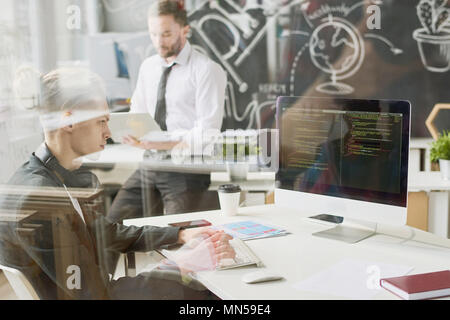 Side view portrait of contemporary young man coding using computer sitting at desk and working in startup project, shot from behind glass wall, copy s Stock Photo