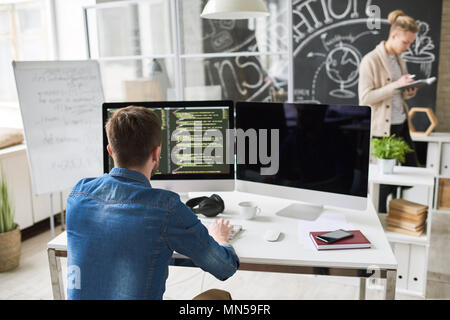 Back view portrait of contemporary program developer writing code sitting at desk with computer and working on startup project in modern office, copy  Stock Photo