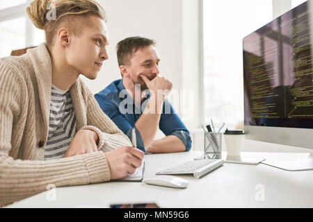 Side view portrait of two contemporary program developers writing code sitting at desk using computer and collaborating on startup project in modern o Stock Photo