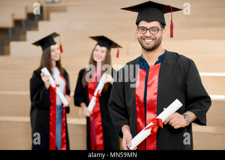 Happy smiling boy keeping university diploma. Graduate standing near young groupmates and looking satisfied. Students wearing black and red colored graduation gowns. Ending university.