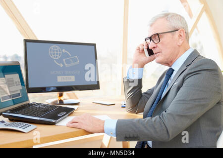 Portrait of senior investment businessman wearing suit while sitting at office desk in front of laptop and making call. Stock Photo