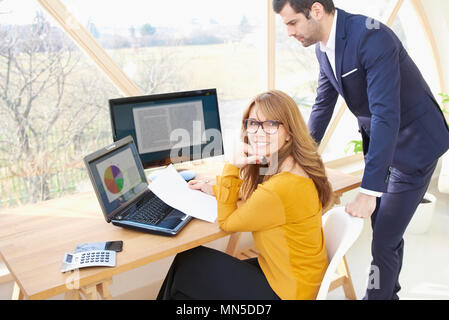 Smiling mature businesswoman sitting at office desk while her young colleague standing next to her and working on financial report together. Teamwork  Stock Photo