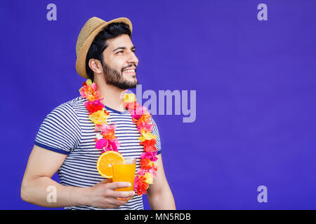 Handsome bearded man dressed in lei on neck and hat and holds orange drink on purple background, vacation time concept, isolated studio shot Stock Photo