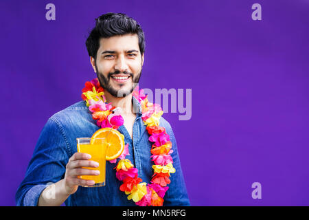 Bearded man dressed in lei on neck holds orange drink on purple background, vacation time concept, isolated studio shot Stock Photo