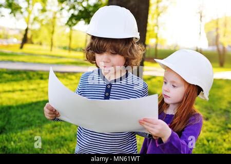 boy and girl in construction helmets looking at white sheet of paper or drawing and smiling Stock Photo