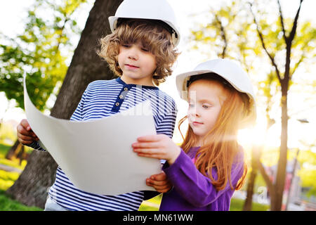 boy and girl in construction helmets looking at white sheet of paper or drawing and smiling Stock Photo