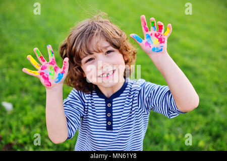 boy with curly hair without front milk tooth shows hands dirty with multi-colored finger paints and smiles. Stock Photo