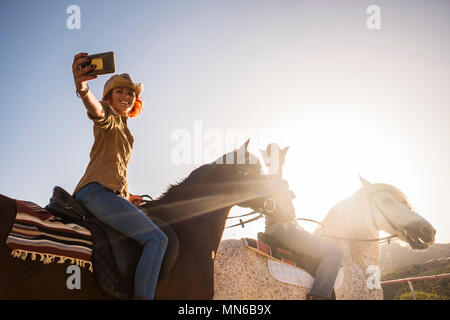 couple on horses riding outdoor under the sunlight in backlight for leisure activity taking selfie with modern mobile phone technology. alternative li Stock Photo