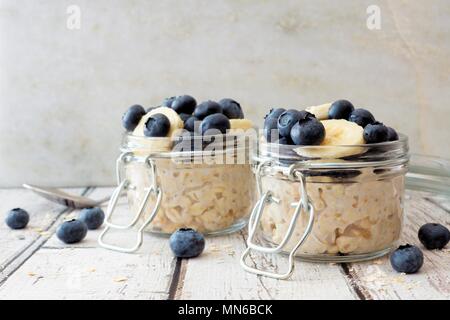 Overnight oats with fresh blueberries and bananas in jars on a rustic white wood background Stock Photo