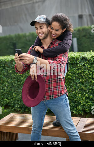 Cheerful interracial couple is posing outdoors. White guy in a red shirt holds a smartphone and his black girlfriend hugs him from behind while stands Stock Photo