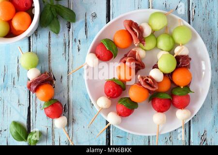 Plate of delicious summer fruit skewers with melon, cheese and prosciutto on a rustic blue wood background Stock Photo