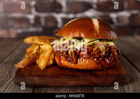 Pulled pork burger on pretzel bun with potato wedges on a wood server with brick background Stock Photo