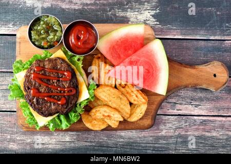 Picnic scene with open hamburger, potato wedges and watermelon on a paddle board over rustic wood Stock Photo
