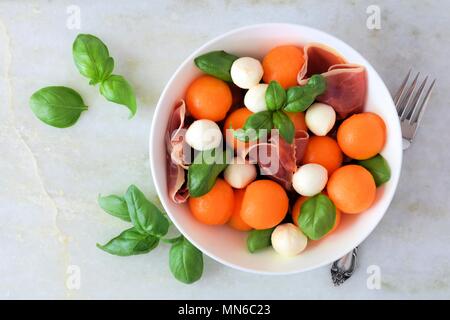 Summer salad with cantaloupe, mozzarella, prosciutto, and basil, above view on a white marble background Stock Photo