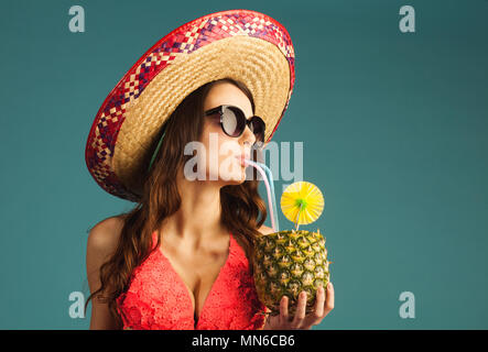 young beautiful woman in bikini, sunglasses and sombrero drinking tropic cocktail isolated on blue background, close-up Stock Photo