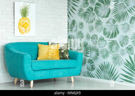 Yellow pillow on blue sofa against a wall with pineapple poster in living room with floral wallpaper Stock Photo
