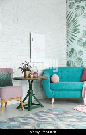Green table with plant between turquoise sofa and pink chair against white brick wall with poster in living room