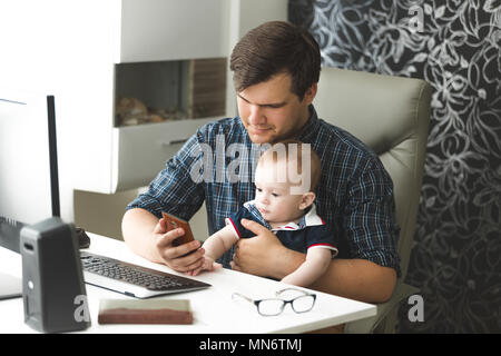 Toned portrait of young selfemployed man working at home with computer and mobile phone and looking after his baby son Stock Photo