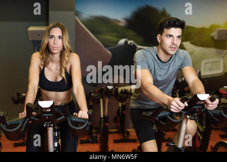 Two people biking in the gym, exercising legs doing cardio workout cycling bikes. Couple in a spinning class wearing sportswear. Stock Photo