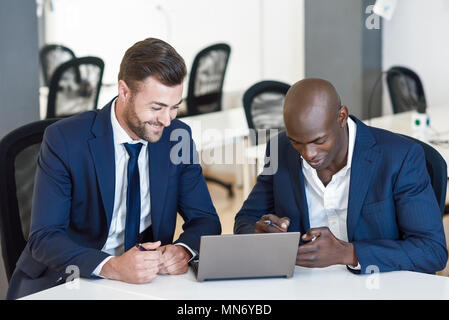 Black and caucasian smiling businessmen looking at a laptop computer. Two men wearing blue suits working in an office with white furniture Stock Photo