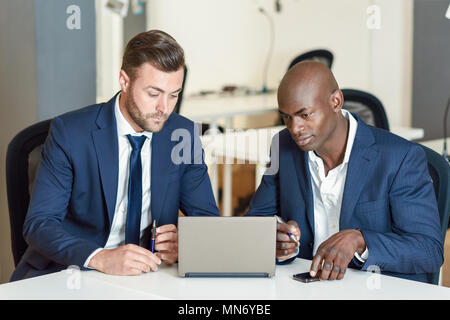 Black and caucasian businessmen looking at a laptop computer. Two men wearing blue suits working in an office with white furniture Stock Photo