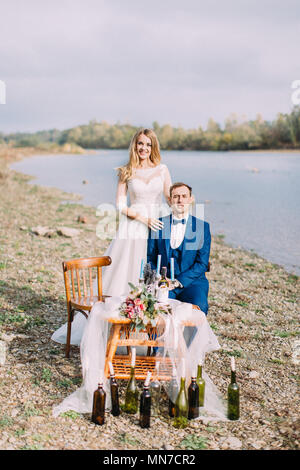 The bride is standing behing the sitting groom at the table placed on the coast. Stock Photo