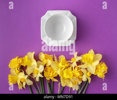 Spring table set with clean elegant porcelain tea or coffee cup and narcissus flowers, vibrant yellow and purple colors, top view, flat lay Stock Photo