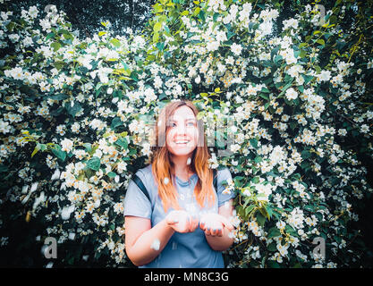 happy young smilling girl having fun catches petals with his hands on background of blooming bush with white flowers of jasmine. Stock Photo