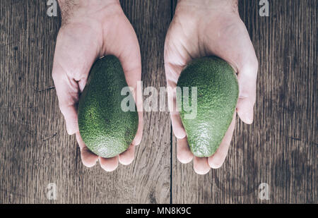 top view of hands of man above rustic wooden table holding two avocados Stock Photo