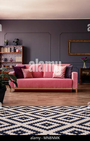 Pink velvet couch with cushions standing in the middle of dark grey living room interior with molding on the wall and geometric carpet Stock Photo