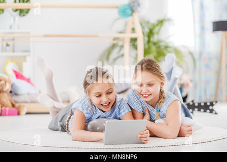 Smiling sisters playing on a tablet while lying on a white rug in a bright room Stock Photo