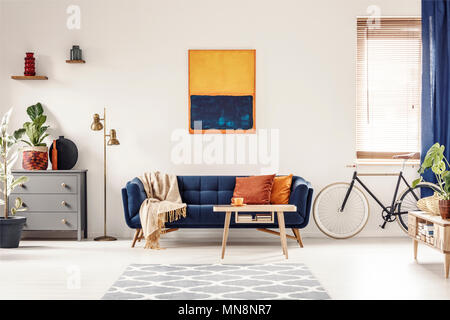 Yellow and blue painting hanging on white wall in bright living room interior with grey cupboard, gold lamp, sofa with blanket and pillows, and bike s Stock Photo