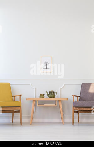 Wooden table between grey and yellow armchair against white wall with poster in living room interior Stock Photo