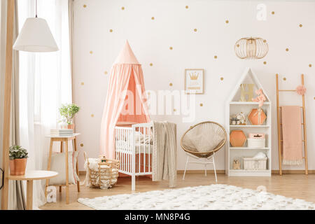 Gold chair with pillow standing next to a white wooden crib with pastel pink canopy in bright baby room interior with dots on the wall Stock Photo