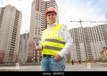 Portrait of smiling male engineer in hardhat standing with blueprints against building under construction Stock Photo