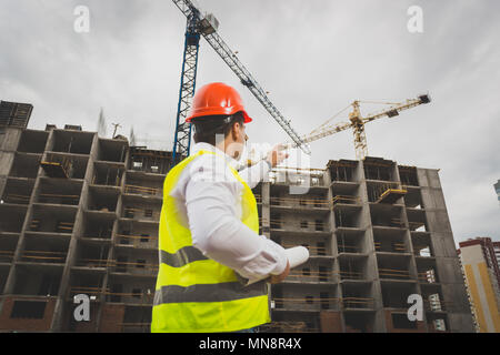 Toned image of male construction worker pointing at working cranes on building Stock Photo