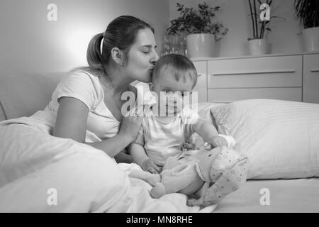 Black and white portrait of young mother kissing her baby son before going to sleep Stock Photo