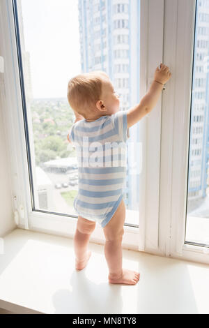 9 months old baby boy standing on windowsil and trying to open the lock on window and pull the handle Stock Photo