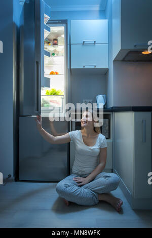 Smiling young woman sitting on kitchen floor at night and opening refrigerator door Stock Photo