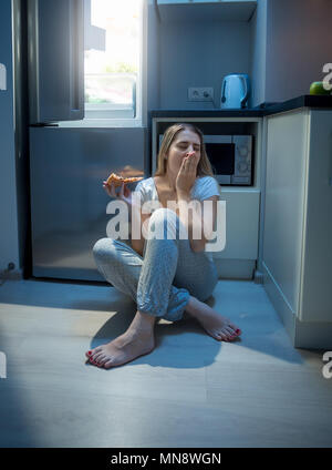 Yougn woman sitting on floor at open refrigerator and eating pizza Stock Photo