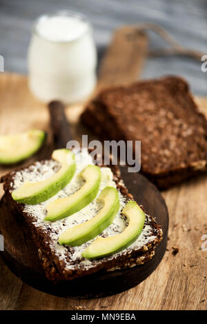 closeup of a slice of rye bread topped with some slices of avocado, and a glass jar of yoghurt, on a rustic wooden table Stock Photo