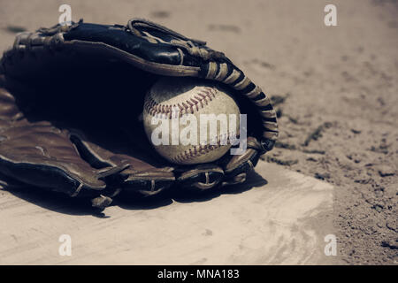 Vintage baseball in players glove laying on homeplate, ball field dirt in background. Stock Photo