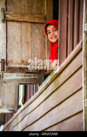 Krabi, Thailand - May 2, 2015: Cute muslim girl wearing red hijap moving out of wooden windows smiling to visitors in rural of Krabi, Thailand Stock Photo