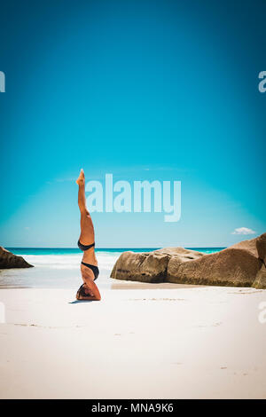 Mid adult woman in bikini doing headstand at beach against clear blue sky during sunny day Stock Photo