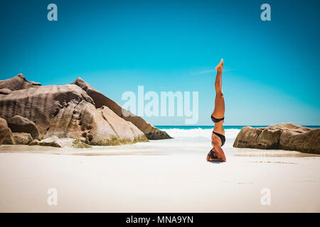 Side view of mid adult woman in bikini doing headstand at beach against clear blue sky, Seychelles Stock Photo