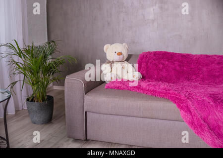 Teddy Bear - sitting on sofa couch with purple blanket, plaid. Palm tree as home plant is near sofa.Broun beige teddy bear, soft toy.Copy space Stock Photo