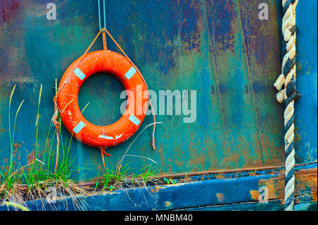 Close up of a life buoy hanging over the side of a beached steel ship.  Rope and buoy contrast against colorful rusting metal hull as grass grows from Stock Photo